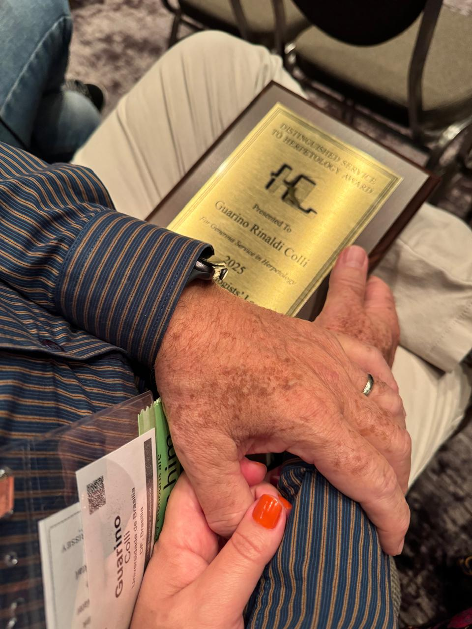 Close-up da placa do prêmio e da mão de uma mulher segurando o braço do professor.